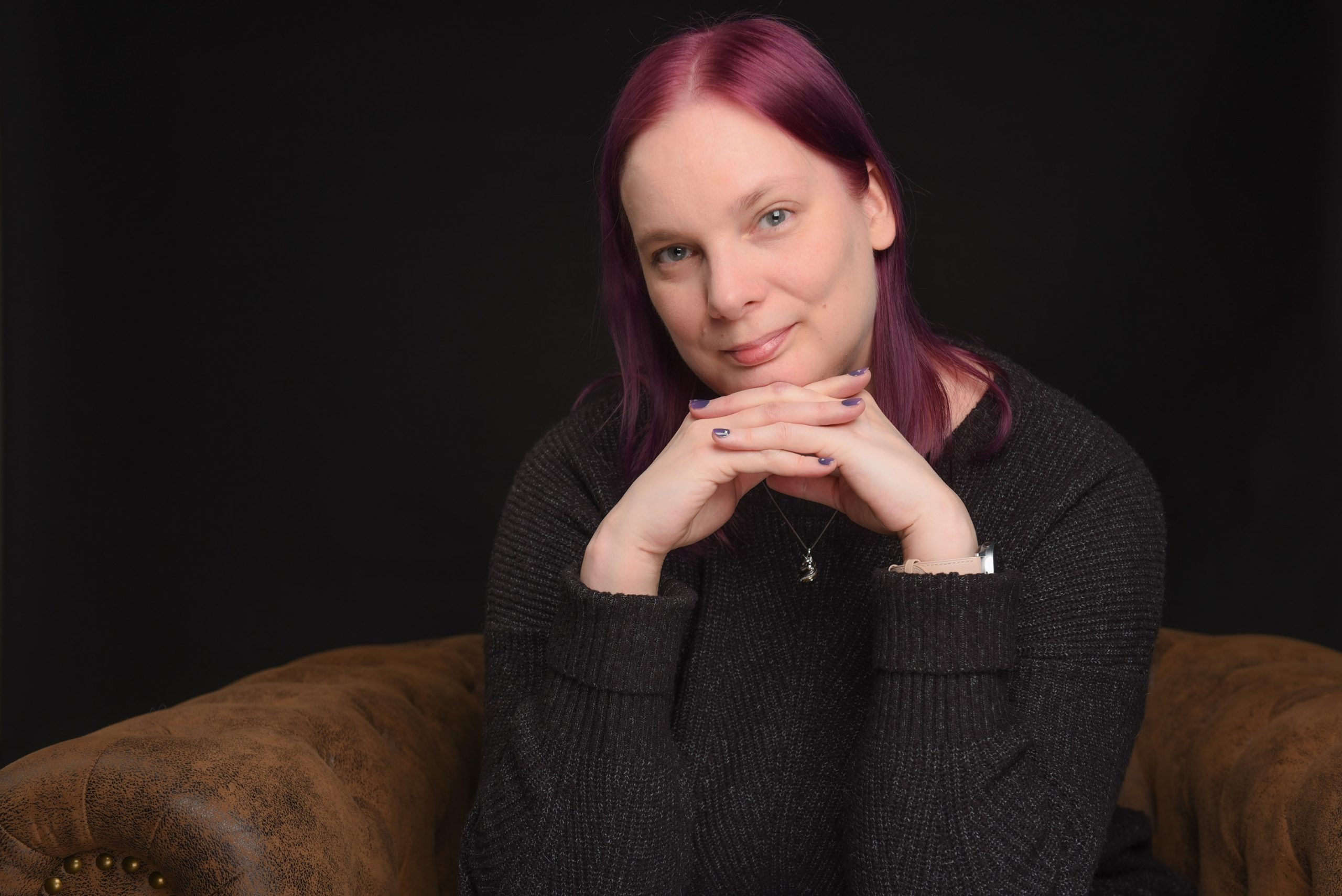 Nadine Most mit lila Haaren sitzt auf einem braunen Sessel und stützt das Kinn auf verschränkten Fingern ab